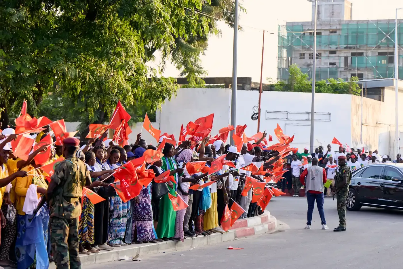 Foule alignée le long d'une avenue, agitant des drapeaux rouges en fin de journée.