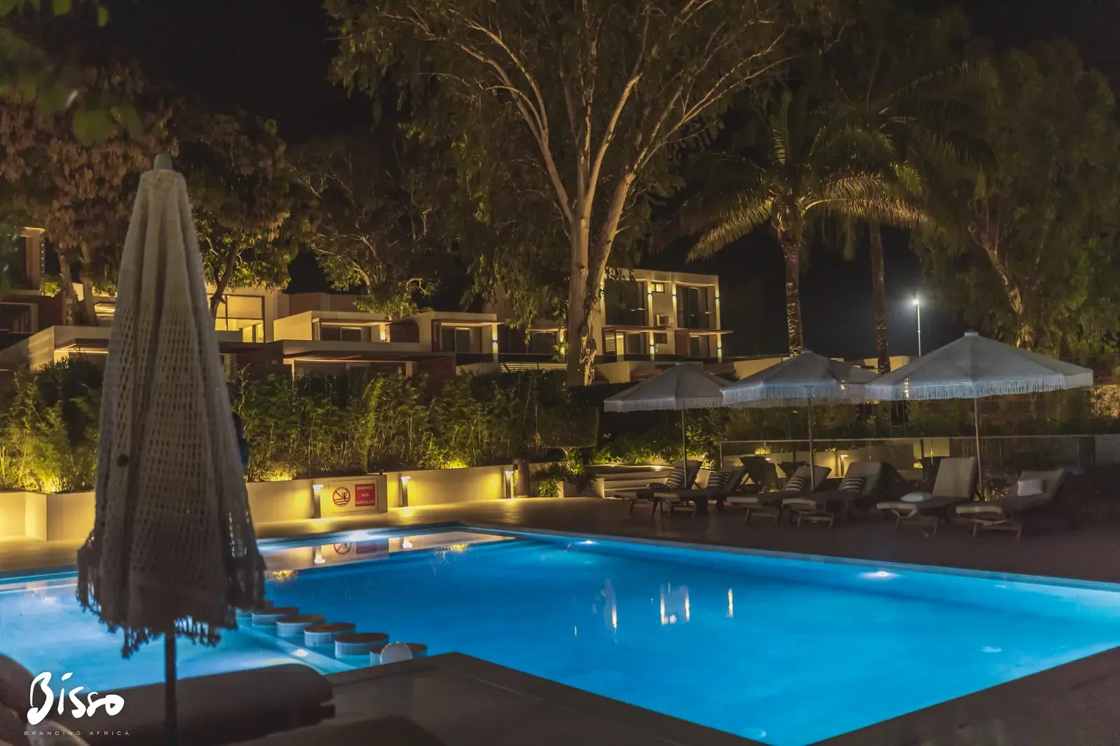 La piscine éclairée en bleu la nuit, avec ses transats, ses parasols et les bâtiments de la résidence en arrière-plan.
